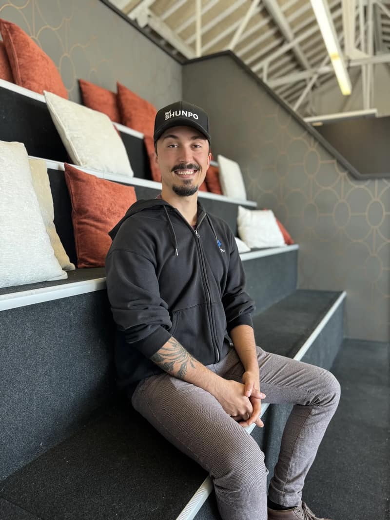 Un membre de l'équipe SHUNPO assis sur les gradins du bureau, casquette SHUNPO sur la tête, souriant.