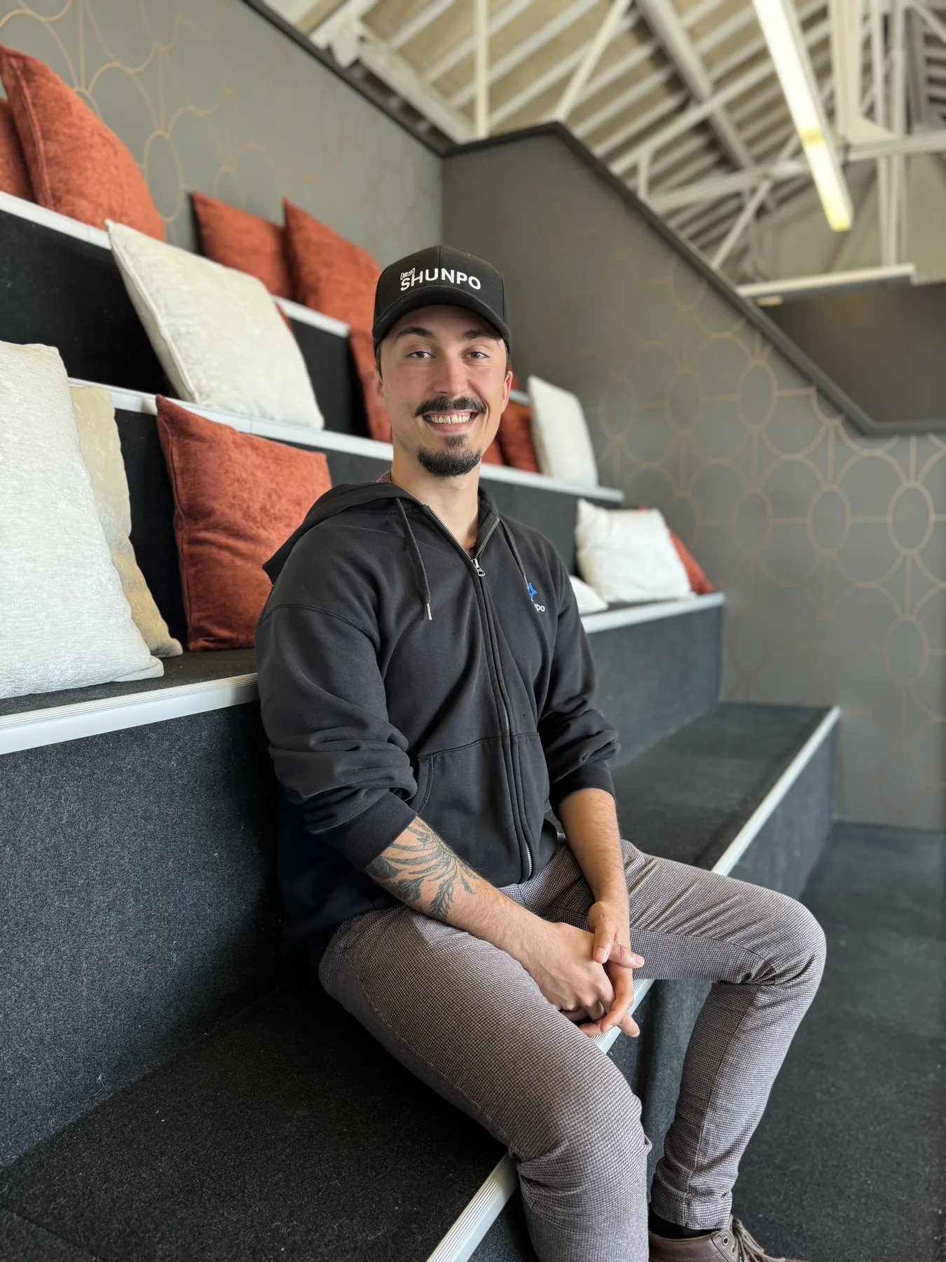 Un membre de l'équipe SHUNPO assis sur les gradins du bureau, casquette SHUNPO sur la tête, souriant.