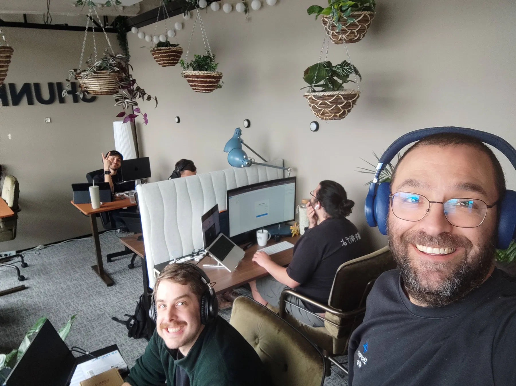 Selfie d'un membre de l'équipe SHUNPO, casque bleu sur les oreilles, avec ses collègues au travail derrière lui.